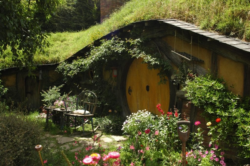 Yellow hobbit house with a chair out the front covered in flowers and foliage